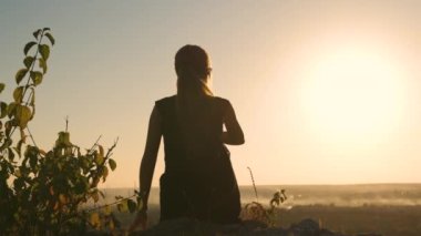 Siyah kısa elbiseli genç, sıska bir kadın yaz günbatımında bir kayanın üzerinde oturmuş dinleniyor. Modaya uygun bir kadın doğada sıcak bir akşamın tadını çıkarıyor..