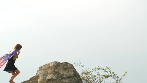 年轻快乐的女人 长发飘扬 随风飘扬 手中拿着美国国旗 站在高高的岩石山上 享受夏日的温暖 — 图库视频影像
