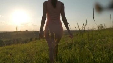 Pembe yaz elbiseli genç bir kadın gün batımında yeşil alanda yürüyor..