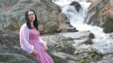 Uzun, şık bir gece elbisesi giymiş, siyah saçlı bir kadın hızlı akan suyla birlikte küçük bir dağ nehrinin kıyısındaki bir taşın üzerinde oturuyor..