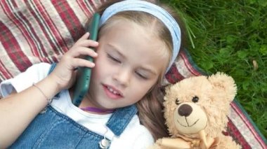 Küçük mutlu kız çocuğu yazın yeşil çimlerde battaniyeye uzanmış oyuncak ayısıyla cep telefonuyla konuşuyor..