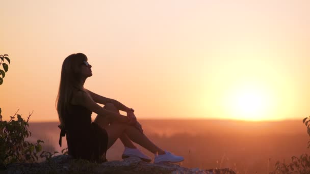 穿着黑色短短的夏装的年轻漂亮女人在日落时坐在岩石上放松 时尚女性享受大自然温暖的夜晚 — 图库视频影像