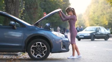 Kafası karışmış bir kadın sürücü arabasının yanındaki caddede durmuş kaputu patlamış ve bozuk motora bakıyor..