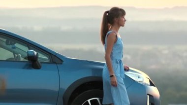 Mavi elbiseli mutlu genç kadın sürücü sıcak yaz akşamının tadını çıkarıyor arabasının yanında duruyor. Seyahat ve tatil kavramı.