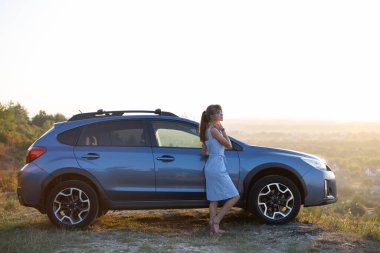 Mavi yazlık elbiseli mutlu kadın sürücü arabasının yanında sıcak bir akşamın tadını çıkarıyor. Seyahat ve tatil kavramı.