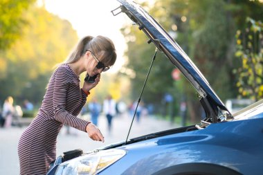 Kaputu açık, bozuk bir arabanın yanında cep telefonuyla konuşan genç üzgün bir kadın sürücü şehir caddesinde aracıyla sorun yaşaması için yardım bekliyor..