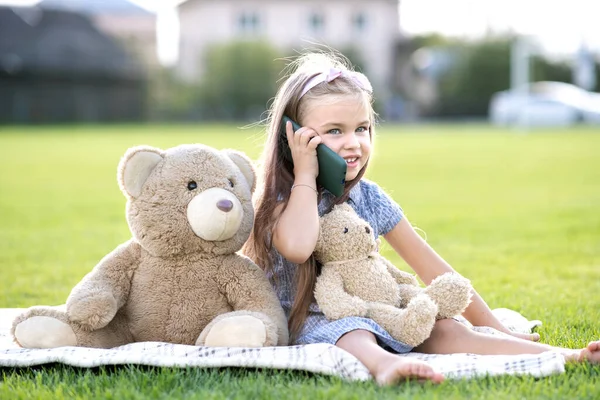 可爱的小女孩与玩具熊在公园的绿草草坪上休息 她的玩具玩具玩具玩具在室外用手机交谈 — 图库照片