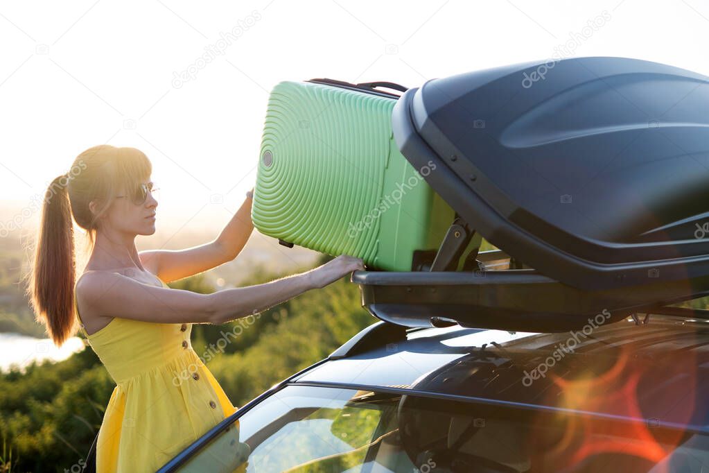Young woman taking green suitcase out from car roof rack. Travel and vacations concept.