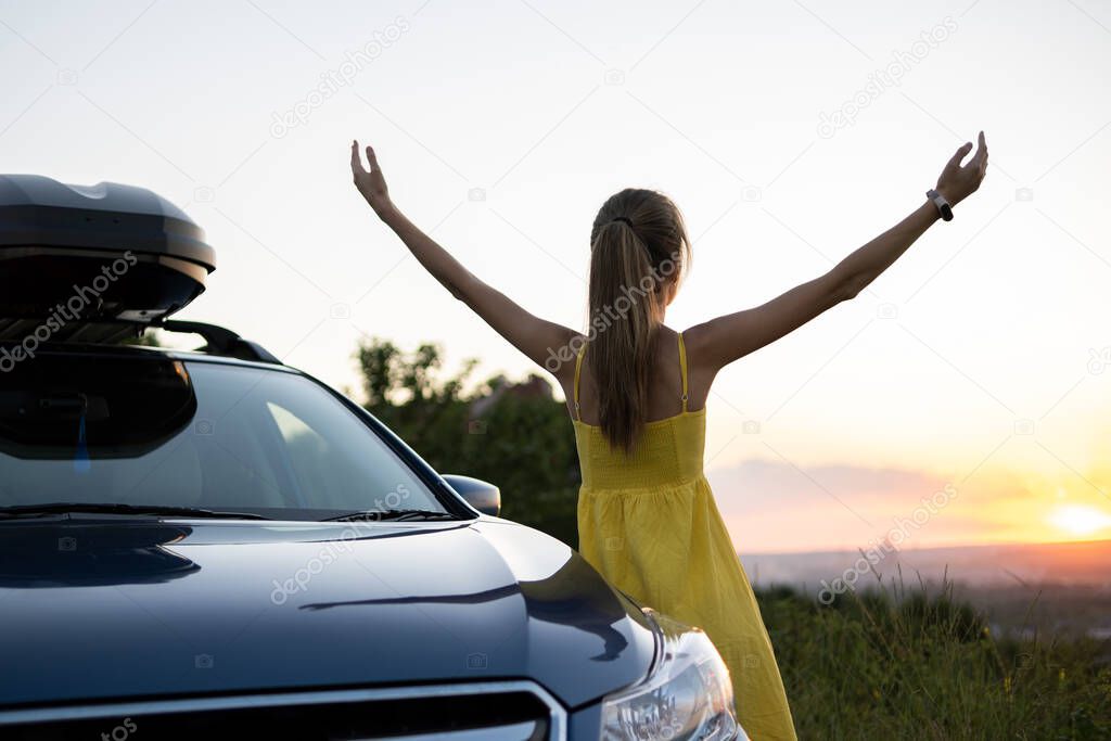 Happy young woman driver in yellow dress enjoying warm summer evening leaning on her car.