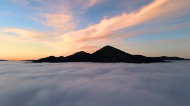 Parlak gündoğumunda beyaz yoğun bulutların üzerinde büyük bir dağın havadan görünüşü.