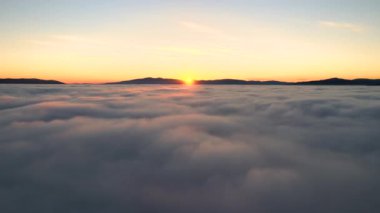 Ufuktaki uzak karanlık dağlarla beyaz yoğun bulutların üzerinde canlı günbatımının havadan görünüşü.