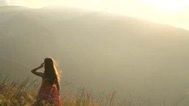 Kırmızılı genç kadın çimenlerin üzerinde duruyor rüzgarlı bir sonbahar akşamında doğa manzarasının tadını çıkarıyor..