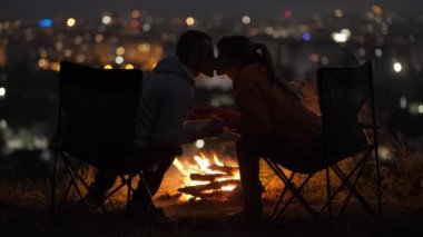 Birbirine aşık genç bir çift gece şenlik ateşinin yanındaki sandalyelerde oturmuş sıcak romantik bir akşamın tadını çıkarıyor..