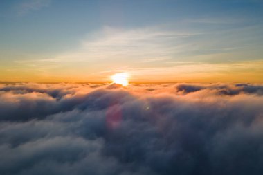 Üzerinde mavi gökyüzü olan beyaz yoğun bulutların üzerinde parlak sarı günbatımının havadan görünüşü.