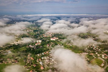 Yazın yeşil tarlalar arasında sıralı binalar ve kıvrımlı sokaklar olan bir kasaba ya da köyün üstündeki beyaz bulutların hava görüntüsü. Yukarıdan kırsal alan.