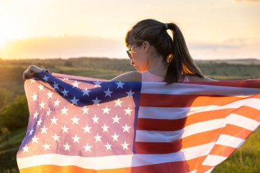Gün batımında ABD bayrağıyla dışarıda dikilen mutlu kadının arka görüntüsü. Amerika 'nın bağımsızlık gününü kutlayan pozitif kız. Uluslararası Demokrasi Günü.