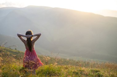 Kırmızılı genç kadın çimenli bir tarlada duruyor rüzgarlı bir sonbahar akşamında doğanın tadını çıkarıyor..