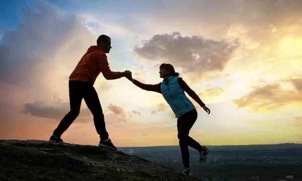 Man and woman hikers helping each other to climb stone at sunset in mountains. Couple climbing on high rock in evening nature. Tourism, traveling and healthy lifestyle concept.