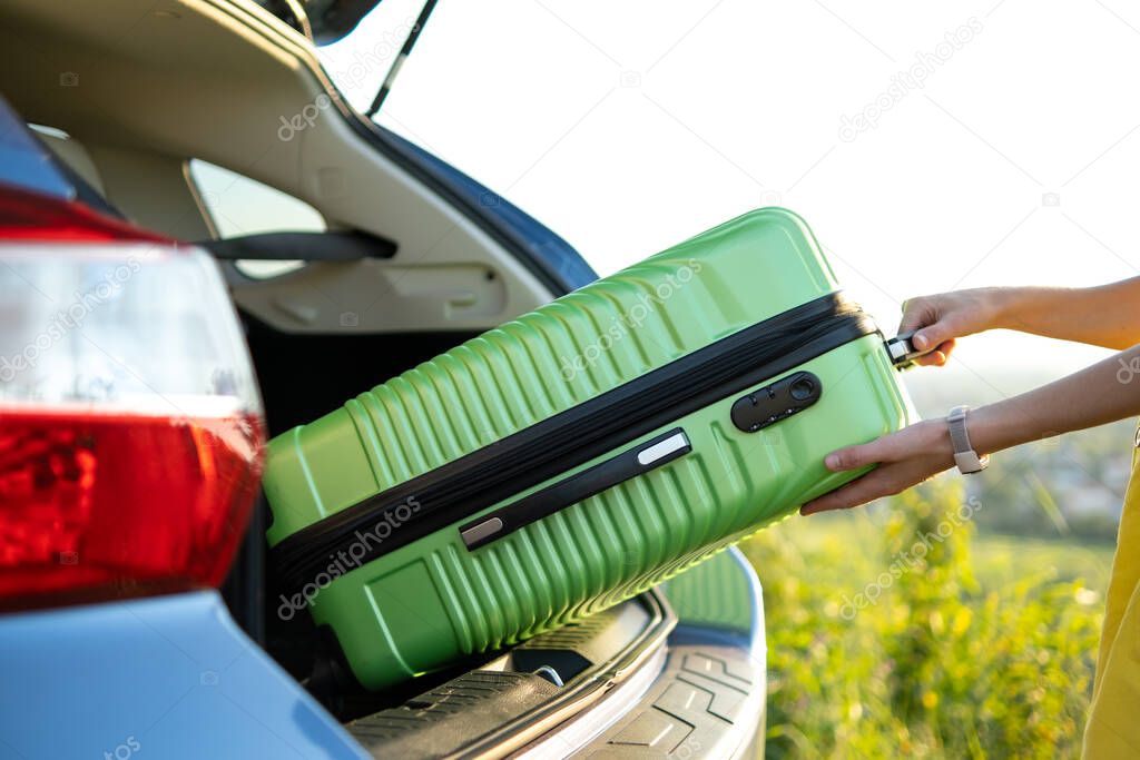 Close up of female hands taking green suitcase from car trunk. Travel and vacations concept.
