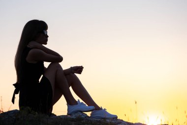 Siyah kısa elbiseli genç, sıska bir kadın yaz günbatımında bir kayanın üzerinde oturmuş dinleniyor. Modaya uygun bir kadın doğada sıcak bir akşamın tadını çıkarıyor..