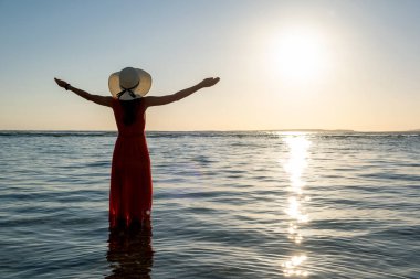 Uzun kırmızı elbise ve hasır şapka giyen genç bir kadın kumsalda deniz suyunda el kaldırarak yaz sabahının erken saatlerinde doğan güneşin tadını çıkarıyor..