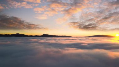 Ufuktaki uzak karanlık dağlarla beyaz yoğun bulutların üzerinde canlı günbatımının havadan görünüşü.