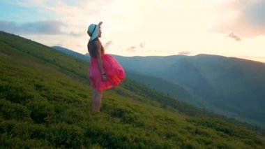 Kırmızı elbiseli genç mutlu kadın gezgin yaz dağlarında rüzgarlı bir akşamda çimenli yamaçta duruyor gün batımında doğanın tadını çıkarıyor..