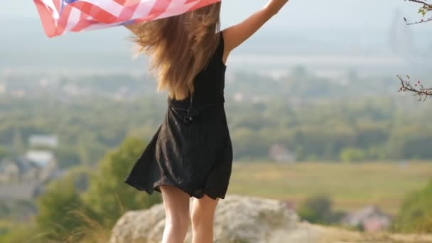 年轻快乐的女人 长发飘扬 随风飘扬 手中拿着美国国旗 爬上高高的岩石山岗 享受夏日的温暖 — 图库视频影像