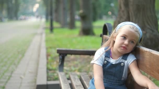 Ένα Μικρό Σοβαρό Κορίτσι Κάθεται Μόνο Του Ένα Παγκάκι Στο — Αρχείο Βίντεο