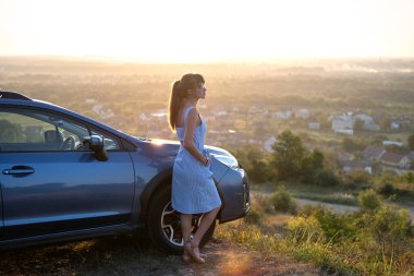 Arabasının yanında dinlenen mutlu kadın sürücü yaz mevsiminin günbatımının tadını çıkarıyor. Seyahat yerleri ve eğlence konsepti.