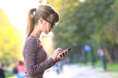 Genç ve güzel bir kadın sıcak bir sonbahar gününde açık bir şehir caddesinde cep telefonuyla mesajlaşıyor..