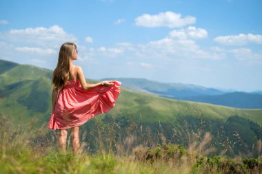 Kırmızılı genç kadın çimenli bir alanda duruyor rüzgarlı bir yaz gününde, dağlarda doğa manzarasının tadını çıkarıyor..