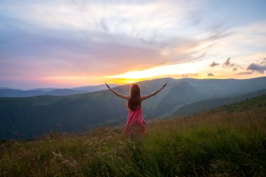 Kırmızılı genç kadın çimenli bir arazide rüzgarlı bir sonbahar akşamında ellerini kaldırarak doğanın tadını çıkarıyor..
