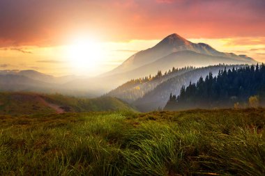 Günbatımı manzarası yeşil çimen çayırı, yüksek tepeler ve kayalık dağlarda canlı akşam gökyüzü altında sisli vadi.