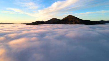 Parlak gündoğumunda beyaz yoğun bulutların üzerinde büyük bir dağın havadan görünüşü.