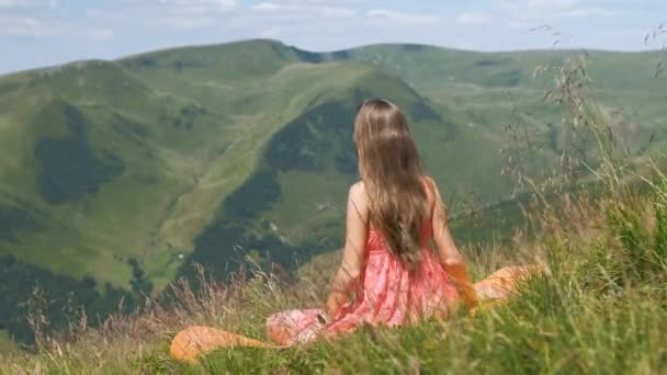 在夏日多风的群山中 身穿红衣的年轻女子坐在草地上欣赏自然风光 — 图库视频影像