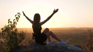 Siyah kısa elbiseli genç, sıska bir kadın yaz günbatımında bir kayanın üzerinde oturmuş dinleniyor. Modaya uygun bir kadın doğada sıcak bir akşamın tadını çıkarıyor..