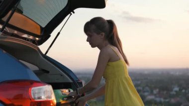 Sarı elbiseli genç kadın arabanın bagajından yeşil bavulu alıyor. Seyahat ve tatil kavramı.