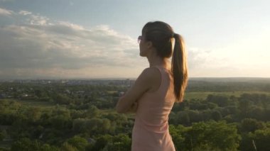 Genç, rahat bir kadın yeşil alanda duruyor ve akşam doğasında gün batımını seyrediyor. Rahatlama ve meditasyon kavramı.