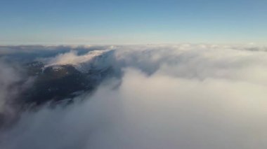 Aydınlık güneşli bir günde karlı dağların tepelerini kaplayan beyaz kabarık bulutların yukarıdan görünüşü.