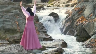Uzun, modaya uygun bir gece elbisesi giymiş genç bir kadın ellerini kaldırıyor ve hızlı akan suyla küçük bir dağ nehrinin kenarında duruyor..