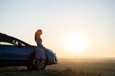 Arabasının yanında dinlenen mutlu kadın sürücü yaz mevsiminin günbatımının tadını çıkarıyor. Seyahat yerleri ve eğlence konsepti.