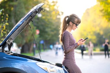 Kaputu açık, bozuk bir arabanın yanında cep telefonuyla konuşan genç üzgün bir kadın sürücü şehir caddesinde aracıyla sorun yaşaması için yardım bekliyor..