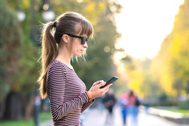 Genç ve güzel bir kadın sıcak bir sonbahar gününde açık bir şehir caddesinde cep telefonuyla mesajlaşıyor..