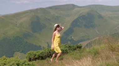 Sarı elbiseli genç kadın yaz dağlarında rüzgarlı bir günde yamaçtaki çayırda yürüyor..