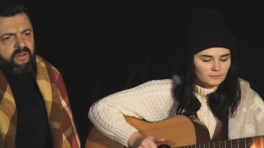 Bir grup insan gece dışarıda şenlik ateşinin yanında oturup gitar çalarak, şarkı söyleyerek ve birlikte mutlu bir şekilde konuşarak eğleniyor..