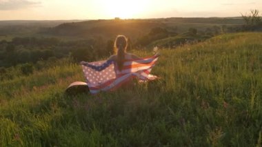 Gün batımında Amerikan bayrağıyla dışarıda oturan mutlu kadının arka görüntüsü. Amerika 'nın bağımsızlık gününü kutlayan pozitif kız. Uluslararası Demokrasi Günü.