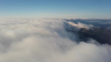 Aydınlık güneşli bir günde karlı dağların tepelerini kaplayan beyaz kabarık bulutların yukarıdan görünüşü.