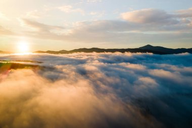 Ufuktaki uzak karanlık dağlarla beyaz yoğun bulutların üzerinde canlı günbatımının havadan görünüşü.
