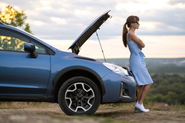 Bozuk bir arabanın yanında duran genç bir kadın sürücü kaputu patlamış ve yardım gelmesini bekliyor..